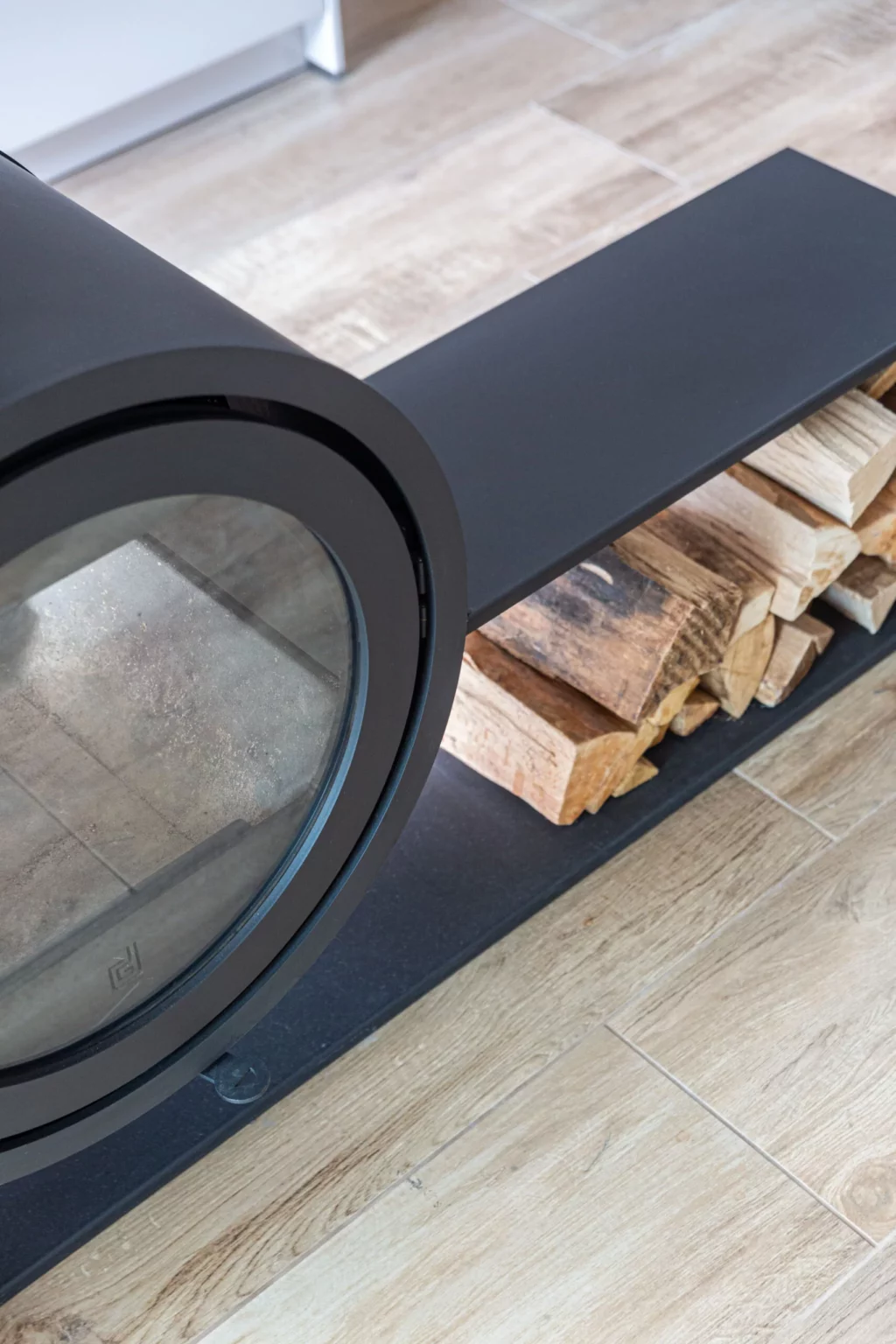 Cheminée design noire suspendue avec espace de rangement pour bûches sur un sol en parquet clair.