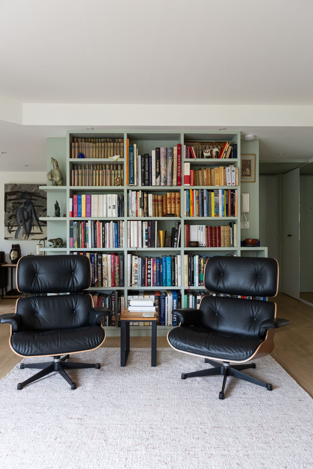 Coin lecture avec fauteuils en cuir noir et bibliothèque sur-mesure couleur vert amande, ambiance cosy et raffinée.