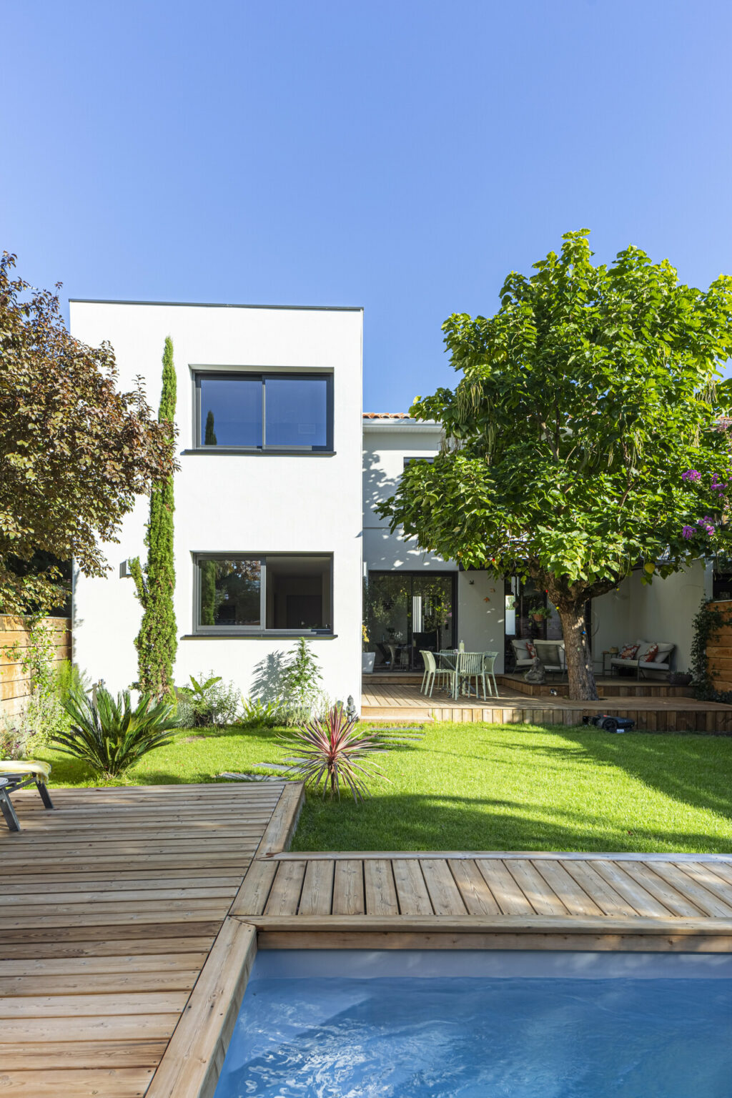 Maison contemporaine avec jardin verdoyant, terrasse en bois et piscine sous un ciel bleu.